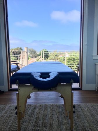 Blue massage table by open sliding doors on a sunny balcony overlooking treetops and a distant coastal ocean view.