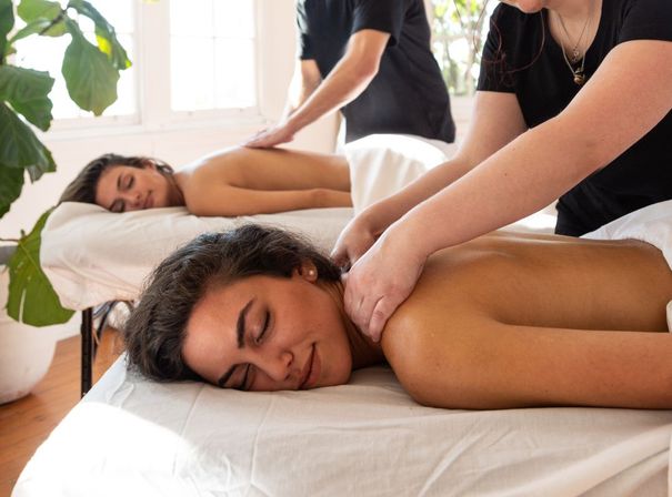Two clients lying face down on massage tables receiving relaxing back massages in a sunlit, plant-filled wellness studio.