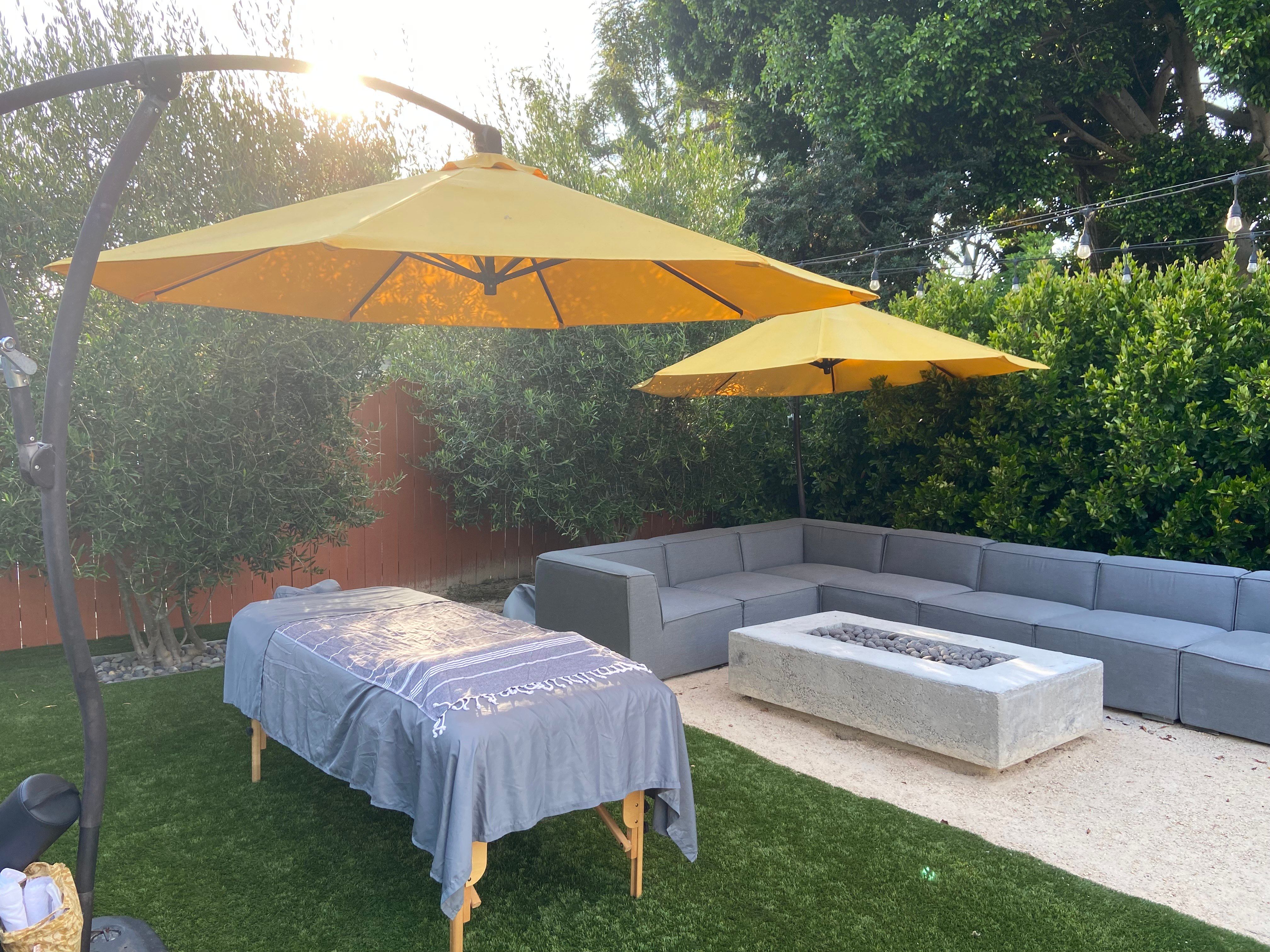 Sunlit backyard patio relaxation setup with two large yellow umbrellas shading a massage table draped in a gray towel, L-shaped gray outdoor sofa, rectangular concrete fire pit, artificial turf and gravel, surrounded by green hedges and string lights.