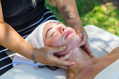 Relaxing outdoor facial: client with towel-wrapped head and gold chain receives gentle jawline massage from an esthetician's hands against a sunlit lawn.