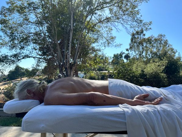 Outdoor massage — person lying face-down on a white-sheeted table with a towel over the lower body, relaxing under sunlit trees and a clear blue sky in a backyard garden setting.