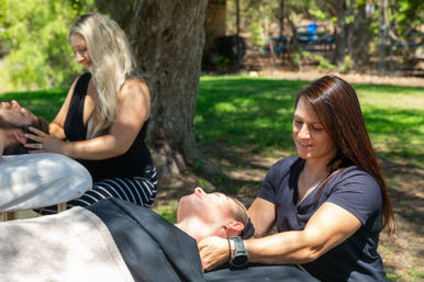 Two massage therapists giving outdoor neck and head massages to clients on portable tables under a shady tree in a sunny park — a serene relaxation and wellness session.