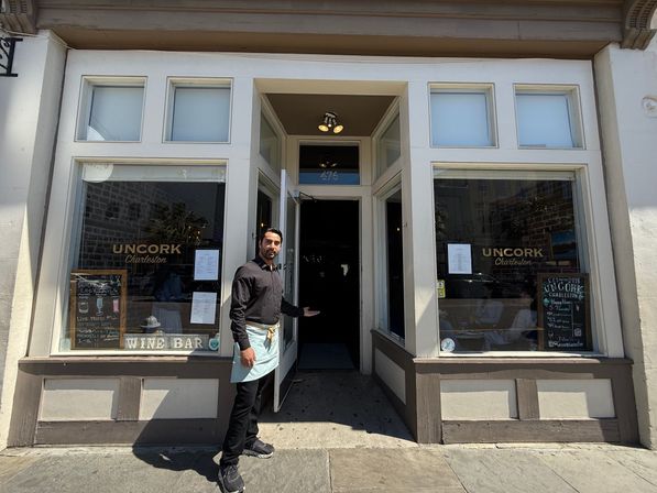 Server in black shirt and apron gestures to the open entrance of a Charleston wine bar storefront with large display windows and sidewalk entrance