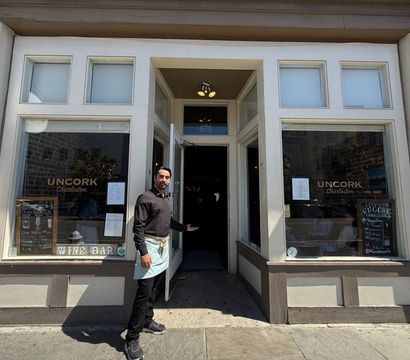 Server in black shirt and apron gestures to the open entrance of a Charleston wine bar storefront with large display windows and sidewalk entrance