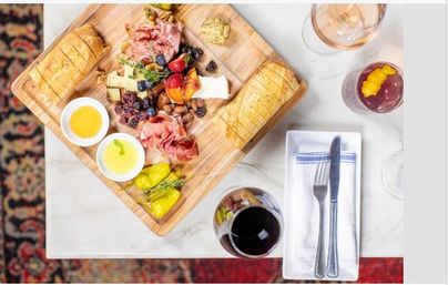 Overhead shot of a wooden charcuterie board with sliced baguette, assorted cheeses, cured meats, berries, nuts and pickled peppers, two dipping bowls, served on a marble table with a glass of red wine, a cocktail and neatly wrapped fork and knife.