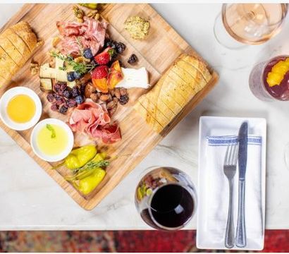 Overhead shot of a wooden charcuterie board with sliced baguette, assorted cheeses, cured meats, berries, nuts and pickled peppers, two dipping bowls, served on a marble table with a glass of red wine, a cocktail and neatly wrapped fork and knife.
