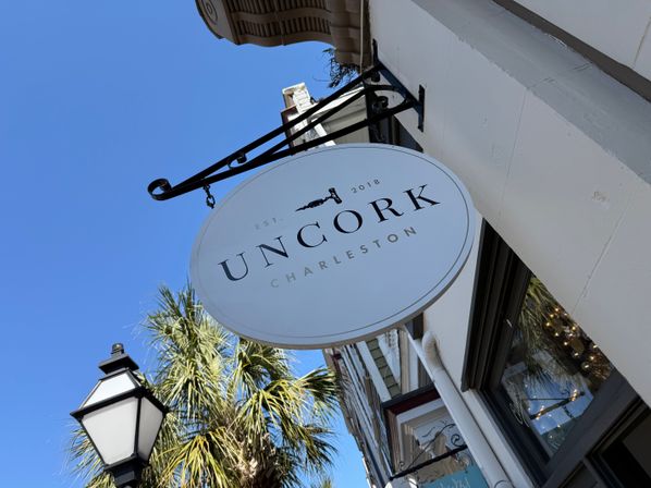 Round white hanging storefront sign on a wrought-iron bracket above a historic Charleston street, palm tree and vintage gas lamp against a bright blue sky