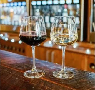 Two stemmed wine glasses side-by-side on a rustic wooden bar—one red, one white—with a blurred wine-dispensing wall and warm lights in a cozy wine bar tasting room.