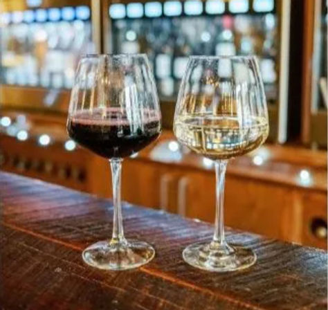 Two stemmed wine glasses side-by-side on a rustic wooden bar—one red, one white—with a blurred wine-dispensing wall and warm lights in a cozy wine bar tasting room.