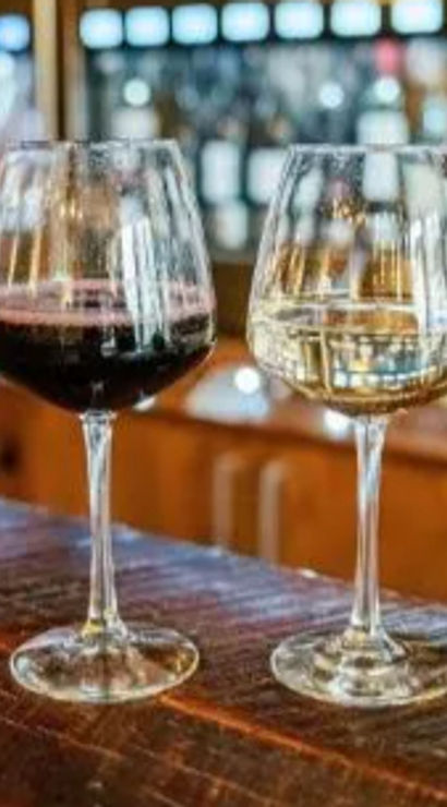 Two stemmed wine glasses side-by-side on a rustic wooden bar—one red, one white—with a blurred wine-dispensing wall and warm lights in a cozy wine bar tasting room.