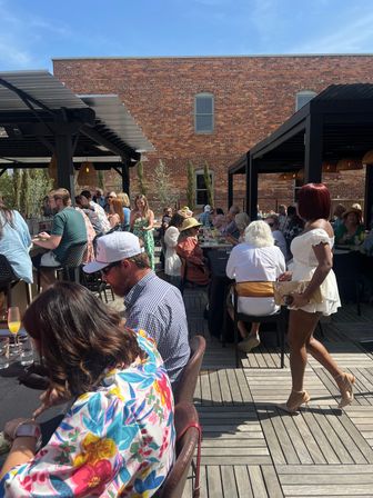 Sunny urban rooftop patio brunch with outdoor dining under pergolas, wooden deck and brick building backdrop, lively crowd enjoying mimosas and summer outfits.