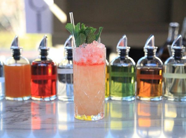 Tall pink-orange cocktail in a highball glass topped with crushed ice, mint sprig and a straw on a marble bar counter, colorful syrup bottles blurred in the background.