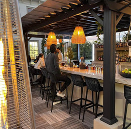 Rooftop patio bar at dusk with marble counter, woven rattan pendant lights, outdoor heater, bottles and patrons seated on black bar stools