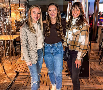 Three smiling women in casual fall outfits—jackets, jeans and boots—posing on a wood outdoor restaurant patio with patio heaters and clear plastic windbreaks at night.