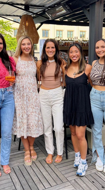 Five women smiling and posing on an outdoor city rooftop patio, wearing casual summer outfits with jeans and sundresses — one holding a cocktail; wooden deck, hanging wicker pendant lights and a bar in the background.