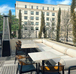 Sunny urban rooftop patio with beige sectional sofa, marble-top dining tables, amber acrylic chairs, tall patio heater, potted cypress trees and mid-rise building backdrop.