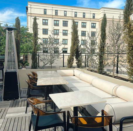 Rooftop patio dining with marble tables, beige cushioned sectional, amber acrylic chairs, tall potted cypress trees and a patio heater against a city building backdrop.