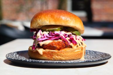Crispy fried chicken sandwich on a glossy brioche bun topped with purple cabbage slaw, sliced pickles and creamy sauce drizzle, served on a patterned plate outdoors.
