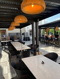 Sunlit urban outdoor patio under a pergola with woven rattan pendant lights, marble-top dining tables, black chairs and a bar with people in the background.