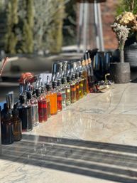 Sunlit patio bar with a neat row of pour‑spout bottles holding colorful syrups and liqueurs on a polished marble countertop, cocktail tools and blurred plants in the background.