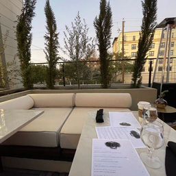 Cozy urban rooftop patio at dusk with cream cushioned banquette, marble table topped with menus, water glasses and coffee cup, and tall slender trees framing city buildings.