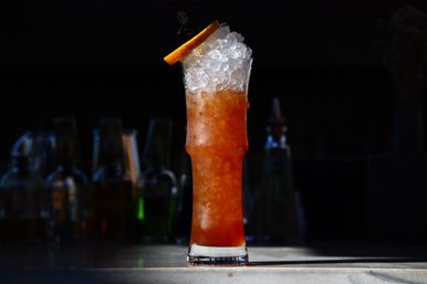 Tall iced amber cocktail in a highball glass topped with crushed ice and an orange slice, sitting on a marble bar counter with blurred liquor bottles in the dark background