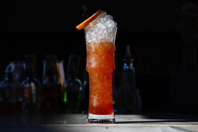 Tall iced amber cocktail in a highball glass topped with crushed ice and an orange slice, sitting on a marble bar counter with blurred liquor bottles in the dark background