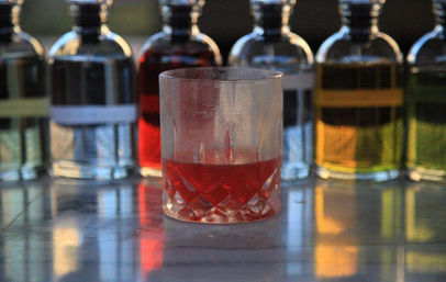 Crystal tumbler with a red cocktail on a reflective marble countertop, vibrant row of colorful liqueur bottles blurred in the background — stylish bar scene.