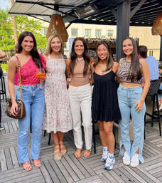 Five friends in summer outfits smiling and posing on an urban rooftop bar patio with wood decking, woven pendant lights, cocktails and city buildings in the background