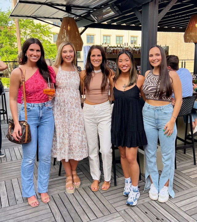 Five friends in summer outfits smiling and posing on an urban rooftop bar patio with wood decking, woven pendant lights, cocktails and city buildings in the background