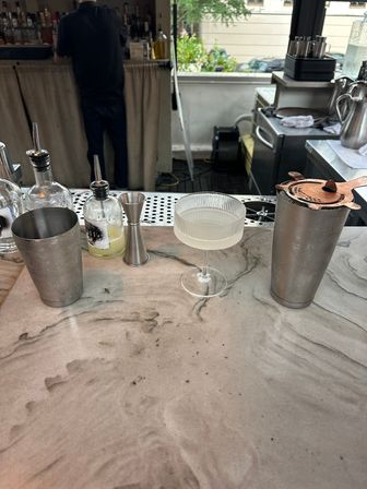 Urban cocktail bar counter with a chilled coupe cocktail, metal shakers, jigger and syrup/bitters bottles on a marbled countertop, bartender working behind the bar and a street view outside.