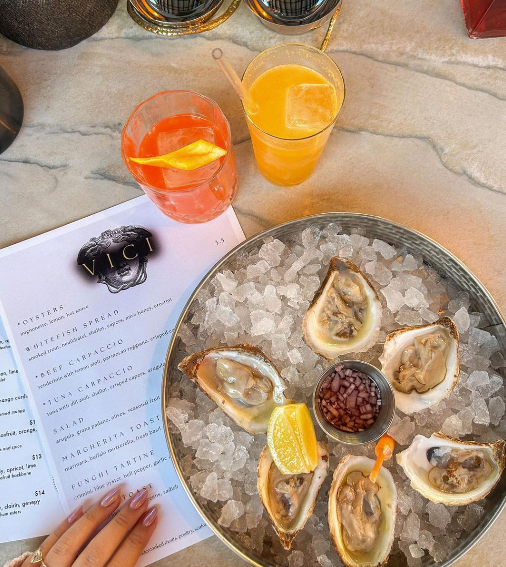 Top-down oyster bar scene: six fresh oysters on crushed ice in a metal platter with lemon wedge and mignonette sauce, two bright cocktails on a marble table and a manicured hand by a menu.