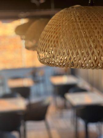 Woven rattan pendant lamp in sharp focus above a cozy cafe seating area with blurred tables, chairs and warm brick wall background