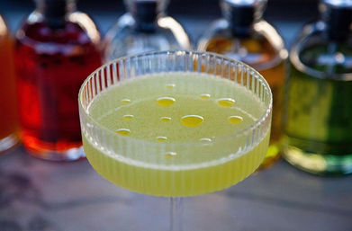 Yellow-green citrus cocktail in a ribbed coupe glass with small oil droplets on the surface, photographed on a bar with colorful bottles blurred in the background