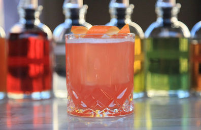 Vibrant pink grapefruit cocktail in a cut-glass rocks glass with orange peel garnish on a bar counter, colorful liquor bottles blurred in the background.