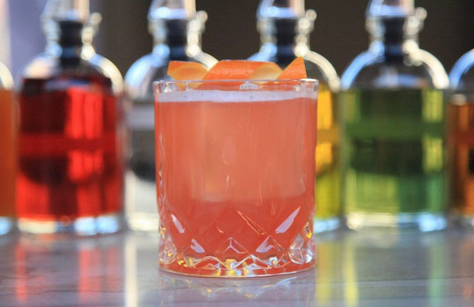 Vibrant pink grapefruit cocktail in a cut-glass rocks glass with orange peel garnish on a bar counter, colorful liquor bottles blurred in the background.