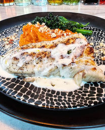 Seared white fish fillet drizzled with creamy sauce, served with sweet potato purée, broccolini and chopped nuts on a stylish black-and-white patterned plate.