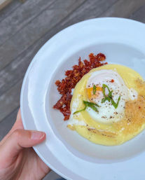 Hand holding white plate with a silky hollandaise-smothered poached egg topped with fresh herbs and cracked pepper, served with crispy bacon bits and a golden parmesan crisp — outdoor brunch on a wooden deck.