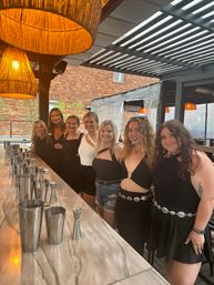 Seven friends in black outfits smiling at a marble-top outdoor patio bar under a metal pergola with rattan pendant lights and a brick wall backdrop, cocktail shakers lined up on the counter.