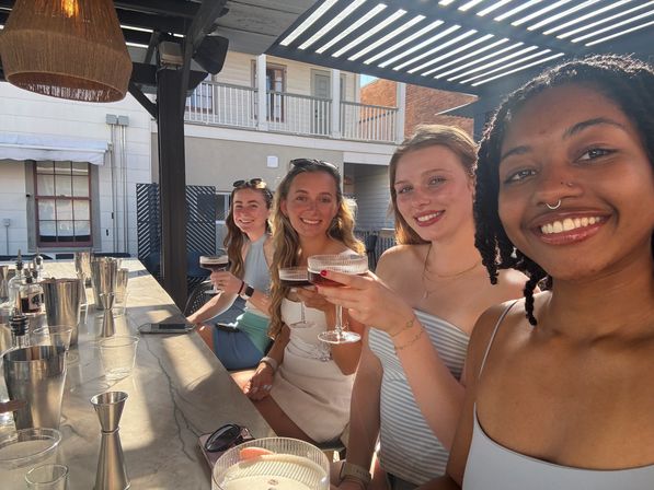 Group of four women smiling and toasting with cocktails at a sunlit outdoor rooftop bar patio under a pergola, seated along a marble counter.
