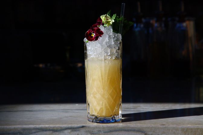 Tall citrus cocktail in a cut-glass highball topped with crushed ice, edible flowers, mint and a straw, sitting on a marble bar against a moody dark background