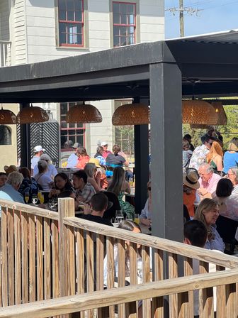 Bustling outdoor restaurant patio on a sunny day with crowds dining and socializing under woven rattan pendant lights, wooden deck railing in foreground and a white building with red-trim windows in the background.
