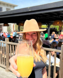 Smiling woman in a tan wide-brim hat holding up a mimosa in a champagne flute for a sunny outdoor brunch toast on a busy patio