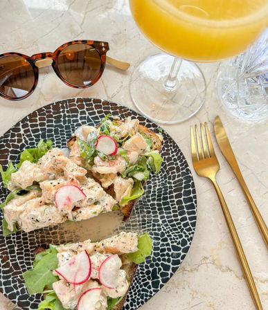Brunch flatlay: creamy salmon salad on open-faced toast with sliced radish and greens on a black patterned plate, gold cutlery and orange mimosa on a marble table with tortoiseshell sunglasses nearby.