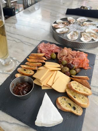 Savory charcuterie board on a marble-topped table: slate platter with sliced smoked cheeses, brie wedge, crostini, fig jam, mixed olives, folded cured meats, plus oysters on ice in a metal tray and a tall glass of white wine.