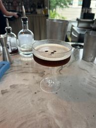 Espresso martini in a ribbed coupe glass topped with three coffee beans, sitting on a marble bar countertop with bottles, jigger and shaker blurred in the background — stylish cocktail bar scene.