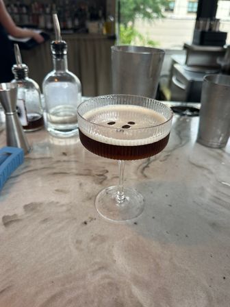 Espresso martini in a ribbed coupe glass topped with three coffee beans, sitting on a marble bar countertop with bottles, jigger and shaker blurred in the background — stylish cocktail bar scene.