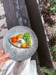 Hand holding patterned plate with colorful avocado toast topped with burrata, red and yellow cherry tomatoes, arugula and pumpkin seeds on a sunny outdoor patio — bright brunch scene