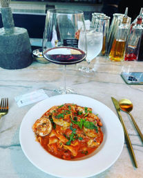 Shrimp and tomato pasta with fresh basil on a white plate at a marble bar counter, paired with a glass of red wine, water, gold cutlery and oil bottles in the background — casual seafood dinner scene.