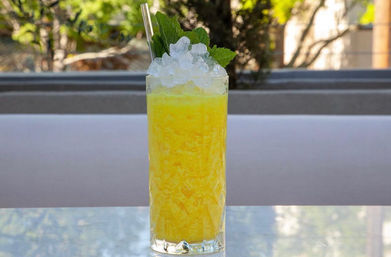 Tall highball glass of bright yellow tropical slushy cocktail topped with crushed ice, fresh mint and a metal straw on a sunlit patio table.